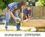 Mid adult carpenter looking at coworker while assisting him in cutting wood with handheld saw - stock photo