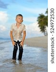 Small photo of Full growth portrait on a tropical beach: handsome and cute 8 years old boy in wet clothes without shoos ankle-deep in water.