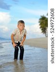 Small photo of Full growth portrait on a tropical beach: handsome 8 years old boy in wet clothes without shoos ankle-deep in water. Leaned his hands on legs, ready for a jump