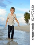 Small photo of Full growth portrait on a tropical beach: handsome 8 years old boy in wet clothes without shoos ankle-deep in water.