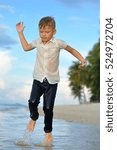 Small photo of Full growth portrait on a tropical beach: handsome 8 years old boy in wet clothes without shoos ankle-deep in water. He is running very fast, splashes falls apart
