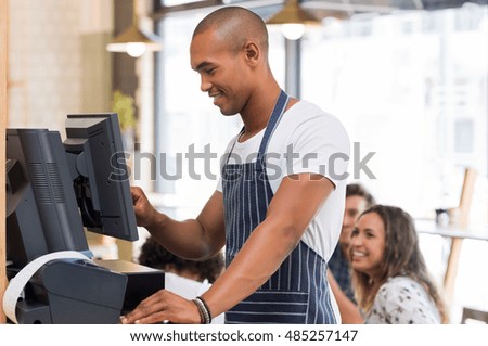 Happy young waiter in blue apron checking bill before printing it. Young african waiter printing bill in cafeteria. Waiter printing order on digital cash register.