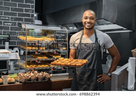 Smiling black baker with apron holding tray of small pastry and looking at camera. Young african chef holding sweet tray at cafeteria. Happy black man smiling at bakery.