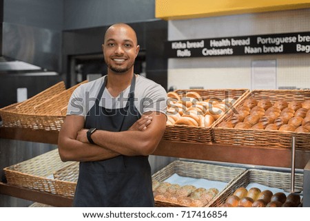Smiling baker man standing with fresh bread at bakery. Happy african man standing with crossed arms at counter in bake shop and looking at camera. Satisfied baker with breads in background.