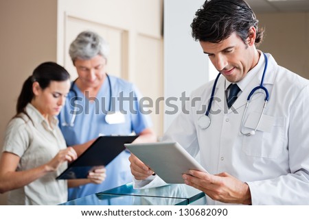 Mid adult male doctor holding digital tablet with colleague and receptionist standing in background
