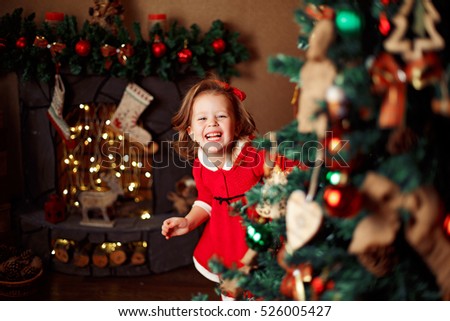 Smiling little girl peeping from behind Christmas tree in living room. Horizontal indoors shot