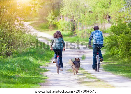 Young happy couple ride bicycles in the village back to camera. Dog running nearby.