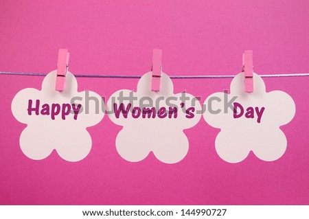Send a Happy Women's Day greeting message on white flower tags hanging from pink pegs on a line against a pink background for International Womens Day, March 8.