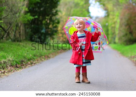 Happy little child, adorable blonde curly toddler girl wearing red duffle coat and holding colorful umbrella walking in the forest or park on a sunny warm spring or autumn day