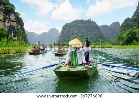 Tourists traveling in boats along the Ngo Dong River at the Tam Coc portion, Ninh Binh Province, Vietnam. Rowers using their feet to propel oars. Landscape formed by karst towers and rice fields.