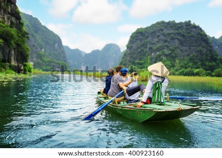 Tourists traveling in boat along the Ngo Dong River and taking picture of the Tam Coc, Ninh Binh, Vietnam. Rower using her feet to propel oars. Landscape formed by karst towers and rice fields.