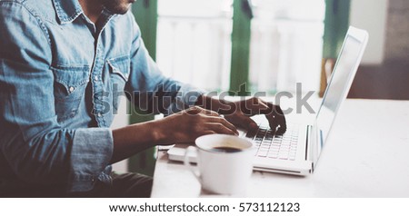 Smiling bearded African man using laptop at home while sitting the wooden table.Male hands typing on the notebook keyboard.Concept of young people work mobile devices.Blurred window background,wide.
