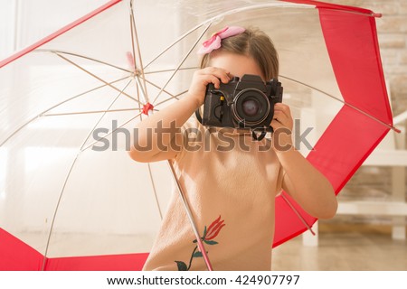 Young little girl photographer with old vintage photo camera taking picture sitting under umbrella, indoor shot. Beautiful girl using vintage photo camera. Children's play. Art or creativity concept.