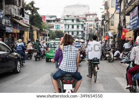 View of a busy street on a weekday in Hanoi, Vietnam. People are seen riding on motorbikes, cycles and cars to their destinations. Market shops are seen on the background.