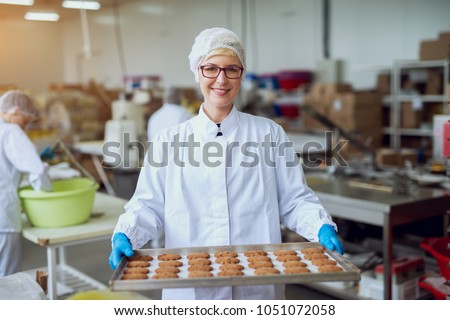 Young happy female worker in sterile cloths holding freshly baked cookies on tinplate inside food production factory.