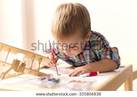 Adorable kid playing on his table with watercolors, painting a wooden parts of his airplane wooden toy. He is young designer with many ideas waiting to be found. Very shallow depth of field.
