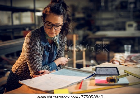 Confident serious attractive professional architect woman leaning against the desk and looking in the new project with a notes, tablet, and rulers on the table in the fabric place.