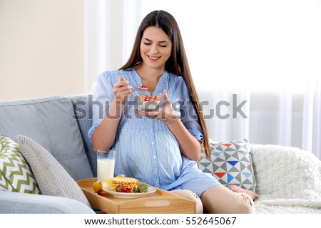 Young pregnant woman with milk and breakfast on sofa