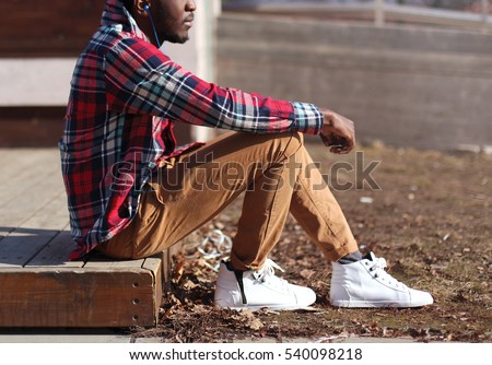 Lifestyle fashion photo stylish african man listens music enjoys sunset, wearing hipster plaid red shirt sitting in profile over evening city background