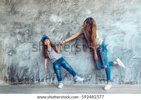 Young beautiful mom with her daughter wearing blank gray t-shirt and jeans posing against rough concrete wall, minimalist street fashion style, family same look, clothing for parent and child.