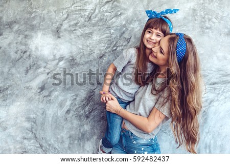 Young beautiful mom with her daughter wearing blank gray t-shirt and jeans posing against rough concrete wall, minimalist street fashion style, family same look, clothing for parent and child.