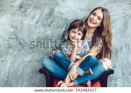 Young beautiful mom with her daughter wearing blank gray t-shirt and jeans posing against rough concrete wall, minimalist street fashion style, family same look, clothing for parent and child.