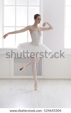 Classical Ballet dancer side view. Beautiful graceful ballerine practice ballet positions in tutu skirt near large window in white light hall. Ballet class training, high-key soft toning.