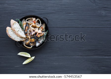 Seafood Stew in Saucepan. Authentic italian restaurant cuisine, healthy food. Oysters, shrimps, calamari in white cream with bruschetta. Bowl on dark black wood background, top view with copy space