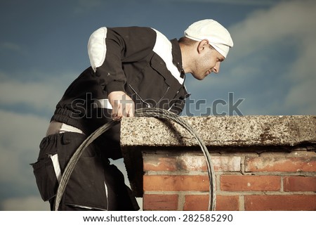 Chimney sweep man in work uniform cleaning chimney on building roof