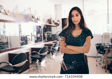 Woman standing with hands folded in her salon. Professional hairdresser with all the hairdressing accessories tied to her waist.