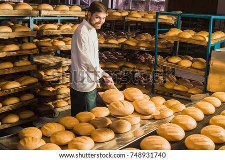 Baker. Bakery. A young handsome worker at a bakery on a background of shelves with spikelets of wheat in his hands. Industrial production of bakery. A man in special clothes of a baker. Working.