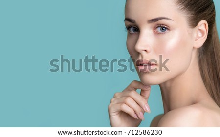 Beautiful young woman with clean perfect skin. Portrait of beauty model with natural nude make up and touching her face. Spa, skincare and wellness. Close up, blue background, copyspace.