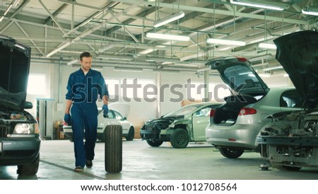 Caucasian young attractive auto mechanic man in blue uniform with blue gloves rolling wheel going on the autoservice. Indoor.