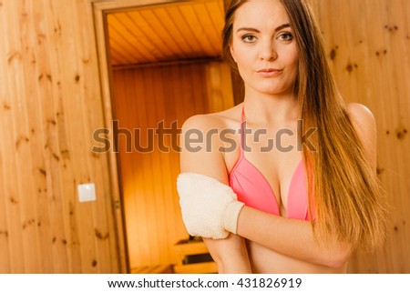 Young woman in wood finnish spa sauna massaging skin with exfoliating glove. Girl in bikini relaxing. Skincare concept.