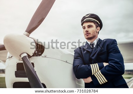 Young handsome man pilot in uniform is standing near small private plane.