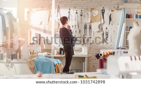 Female fashion, designer, Looking at Drawings and Sketches that are Pinned to the Wall Behind Her Desk. Studio is Sunny. Personal Computer, Colorful Fabrics, Sewing Items are Visible.