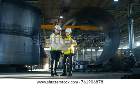 Male and Female Industrial Engineers Look at Project Blueprints While Standing Surround By Pipeline Parts in the Middle of Enormous Heavy Industry Manufacturing Factory