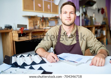 Portrait of smiling young man sitting at table in jeweler shop looking at camera with documentation and ring set in front of him