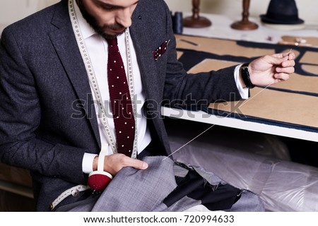 Bearded young tailor wrapped up in sewing male jacket with needle and thread while sitting at working table, interior of atelier on background