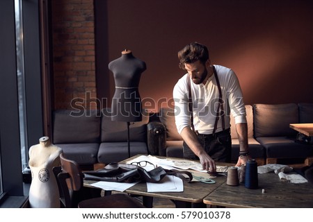Beautiful serious tailor with glasses in white shirt with brown leather suspenders working near wooden table with threads, apron and scissors in amazing atelier with antique furniture and mannequin