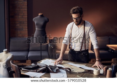 Cute tailor male with beard and glasses in white shirt with brown leather suspenders working near wooden table with threads, apron and scissors in amazing atelier with antique furniture