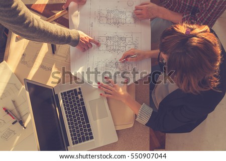 Group of people discussing business plans, with lap-tops, plans in a room lit by natural sunlight.