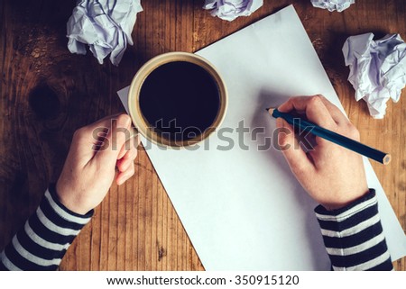 Female writer drinking cup of coffee, top view of female hands holding cup of coffee above pencils and paper on work desk, retro toned image.