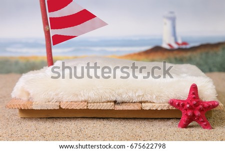 A newborn photography prop of a raft at the beach with sand, a red starfish, a red and white sail and a lighthouse and ocean waves in the blurred background.