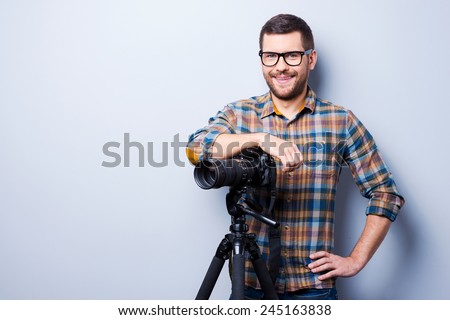 Professional photographer. Portrait of confident young man in shirt holding hand on camera on tripod while standing against grey background