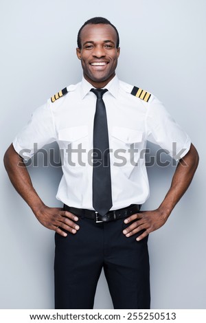 Confident pilot. Confident African pilot in uniform holding hand on hip and smiling while standing against grey background