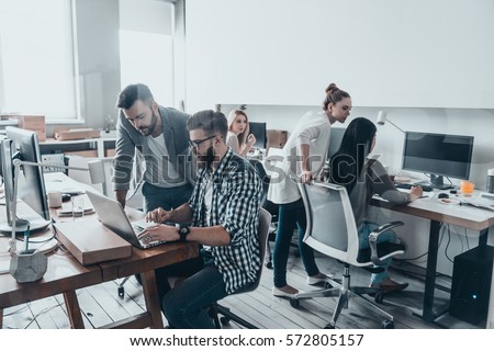 Achieving best results. Two confident young men looking at laptop monitor while their colleagues working in the background