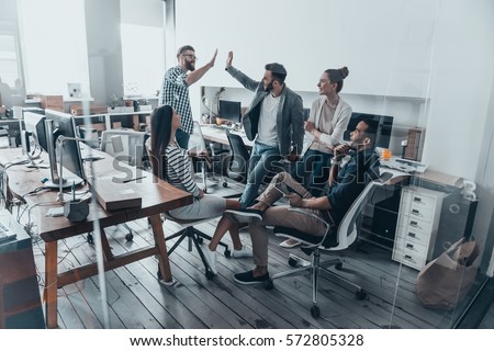 High-five for success! Two cheerful young business people giving high-five while their colleagues looking at them and smiling 