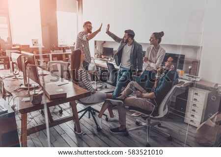 We did it! Two cheerful young business people giving high-five while their colleagues looking at them and smiling 