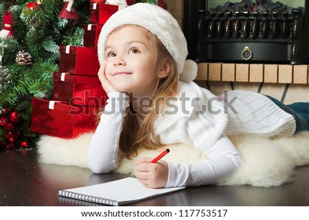 Funny girl in Santa hat writes letter to Santa near christmas tree